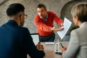 Rücktritt vom Mietvertrag - Ein Mann in einem roten Pullover steht und gestikuliert mit einem Dokument, möglicherweise um einen Rücktritt zu besprechen, in Richtung zweier sitzender Kollegen während einer ernsten Besprechung in einem modernen Büro mit Sichtbacksteinwänden.